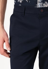 Tamno plave hlače s ravnim prednjim dijelom, kopčanjem na gumbe i bočnim džepovima. Materijal je gladak s laganim sjajem. Minimalistički dizajn.