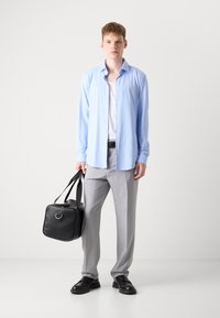Camisa de botones azul claro, pantalones grises, zapatos negros, sosteniendo una bolsa negra con correas. Tela suave, corte relajado, diseño minimalista.