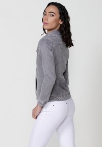 Siva denim jakna s ovratnikom, prilegajoča oblika in stranski razpoki, oblečena čez bele kavbojke. Vidni šivi in podrobnosti kovin na jakni.