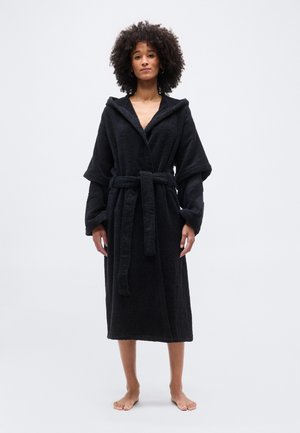 Kobieta o kręconych włosach stojąca boso, ubrana w długi czarny kąpielowy szlafrok z kapturem, związany w pasie, na białym tle.