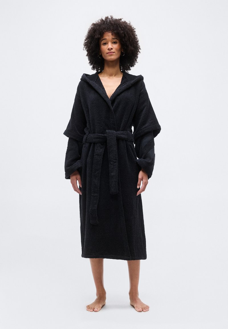 Γυναίκα με σγουρά μαλλιά, ξυπόλητη, φορώντας μακρύ μαύρο μπουρνούζι με κουκούλα δεμένο στη μέση, μπροστά σε λευκό φόντο.