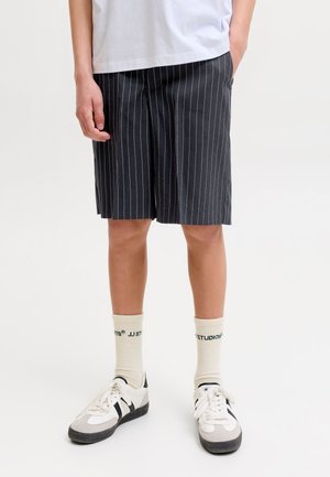 Personne portant un short noir à fines rayures jusqu'aux genoux, des chaussettes blanches avec texte, des baskets blanches et noires à lacets, et un t-shirt blanc.