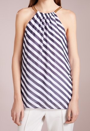 Femme portant un haut en rayures diagonales noir et blanc sans manches avec des bretelles chaînes dorées et un pantalon blanc.