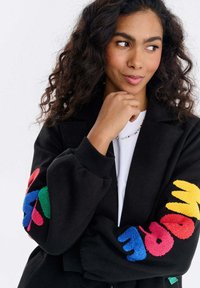 Kobieta o kręconych włosach, ubrana w czarną kurtkę z kolorowymi literami na rękawach, białą koszulę i naszyjnik z koralików, patrząca zamyślona w bok.