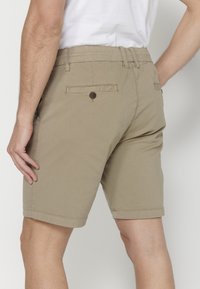 Vīrietis, kas valkā bēšus ceļgalu garuma šortus ar aizmugurējām pogu kabatām un baltu t-kreklu, rādot no aizmugures, krustu augstumā.