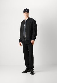 Černá bomber bunda se zipovým zapínáním a žebrovanými rukávy, kombinovaná s černou kravatou, bílou košilí a černými kalhotami a botami. Součástí je také černá čepice.