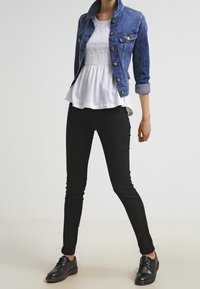 Giacca di denim blu, top peplum bianco, pantaloni skinny neri e scarpe stringate nere. Le caratteristiche includono dettagli con bottoni e maniche arrotolate.
