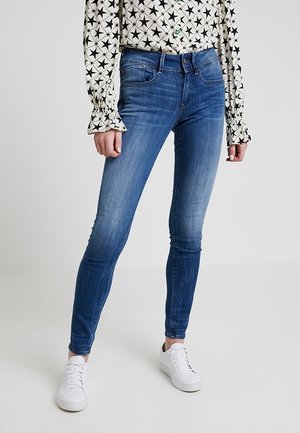 Modré slim fit džíny nošené s bílou halenou s dlouhým rukávem s černými hvězdami a bílými teniskami na světle šedém pozadí.