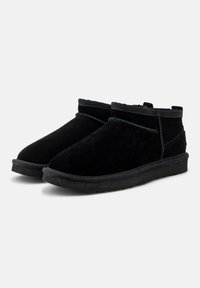 Botas de ante negras hasta el tobillo con un forro suave, punta redondeada y suela de goma plana. Presentan detalles de costura y un puño doblado.