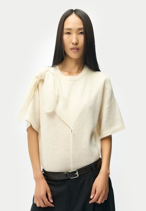 Frau mit langen glatten schwarzen Haaren trägt einen cremefarbenen Kurzarmpullover mit dekorativer Stoffschleife und schwarzem Gürtel über dunklen Hosen.