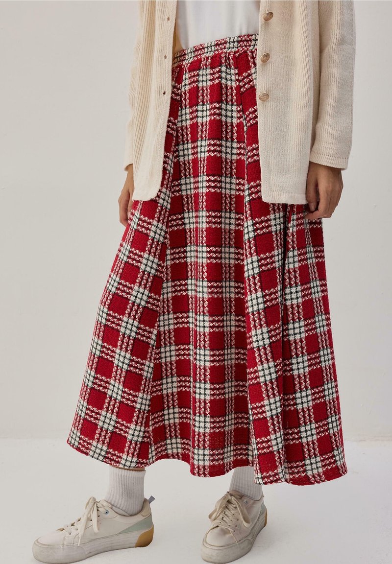 Femme portant une longue jupe à carreaux rouge et blanc, un cardigan beige, des chaussettes blanches et des baskets blanches, debout devant un fond uni.