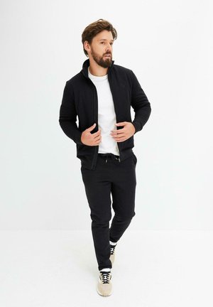 Homme barbu ajustant une veste zippée noire, portant un t-shirt blanc, un jogging noir et des baskets beiges, se tenant contre un fond blanc.