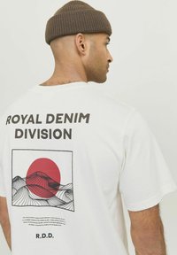 White t-shirt featuring "ROYAL DENIM DIVISION" text, red sun graphic, and wavy line pattern. Short sleeves and relaxed fit.