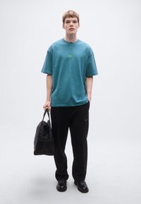 Τυρκουάζ φαρδιά βαμβακερή μπλούζα με μικρό πράσινο λογότυπο, συνδυασμένη με μαύρη φόρμα και κρατώντας μαύρη μαλακή τσάντα duffel.