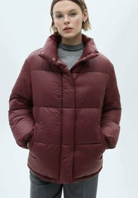 Bordeaux pufferjakke med høj krave, der har et boksformet design, quilted tekstur og trykknapper; vist med en grå turtleneck under.