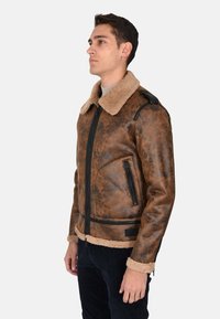 Giubbotto in finta pelle marrone con colletto e polsini in shearling beige. Presenta tasche con zip nere e dettagli in rilievo sul materiale.