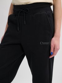 Nahaufnahme schwarzer Jogginghosen mit Kordelzug in der Taille und dem hummel Sportswear-Logo am Oberschenkel, getragen von einer Person mit blauer Nagellackierung.