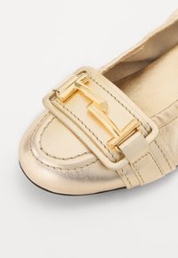 Chaussure plate en cuir doré avec un bout rond, mettant en avant une boucle dorée décorative et un détail de couture sur le dessus. Texture lisse.
