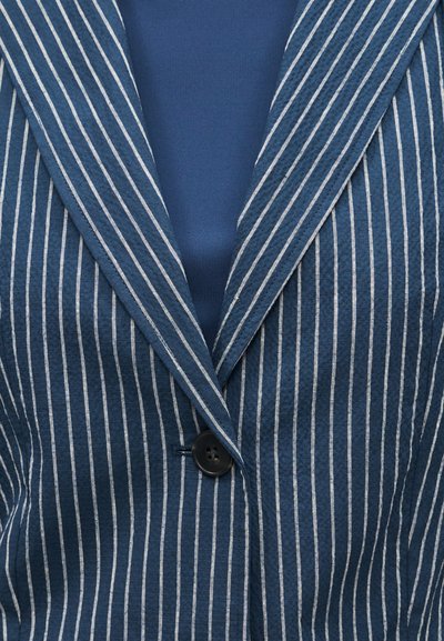 Μπλε σακάκι με στενές λευκές κάθετες ρίγες, ένα μαύρο κουμπί και πέτα με εγκοπές πάνω από ένα μονόχρωμο μπλε πουκάμισο.