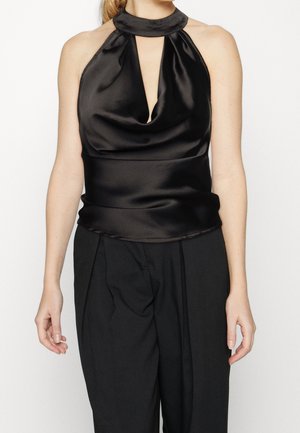 Femme portant un top noir en satin sans manches avec un décolleté en forme de serrure et un pantalon noir ample sur fond blanc.