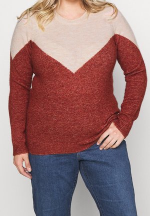 Femme portant un pull à manches longues avec des blocs de couleur crème et rouille et un jean bleu, debout avec une main sur la hanche.