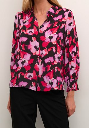 Frau trägt eine langärmelige Bluse mit Knöpfen und einem abstrakten Blumenmuster in Schwarz, Rot und Pink sowie schwarze Hosen.