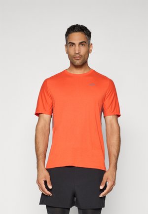 Un homme adulte portant un t-shirt sportif à manches courtes orange vif et un short noir, se tenant de face contre un fond uni.