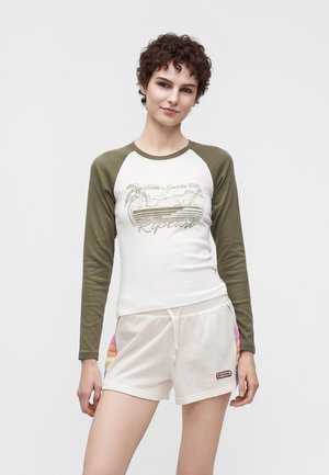 Jong persoon met kort krullend haar, die een wit-groen shirt met lange mouwen en lichte korte broek met Rip Curl-logo draagt, staand tegen een effen achtergrond.