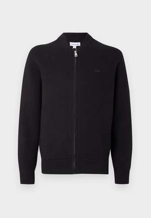 Cardigan zippé noir avec col, poignets et ourlet côtelés, présentant un petit logo brodé sur la poitrine gauche.