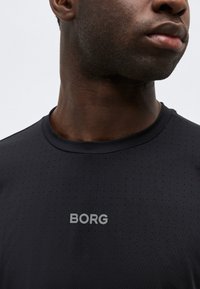 Κοντινό ενός ατόμου που φοράει πουκάμισο μαύρου χρώματος με υφή που παραπέμπει σε πουά και την επιγραφή "BORG" σε γκρι στο στήθος, στραμμένο ελαφρώς προς τα δεξιά.