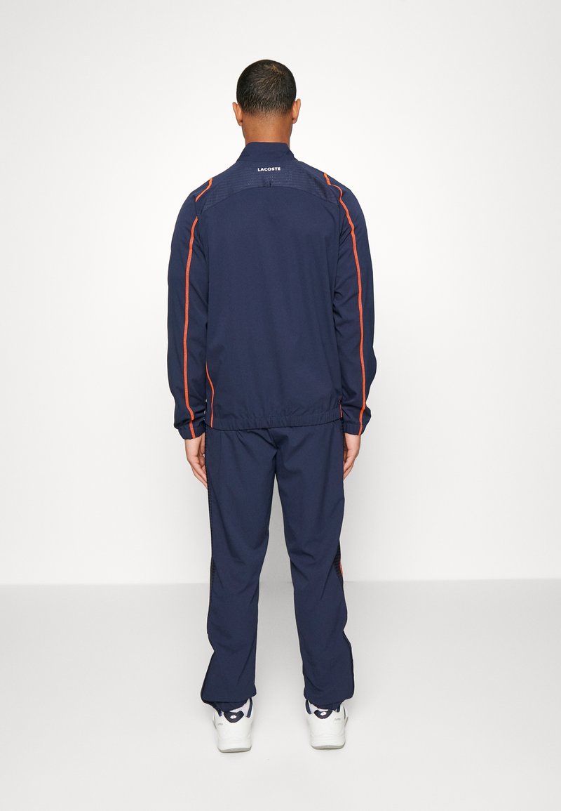 Marineblauwe tracksuit met oranje zijstrepen en elastische tailleband. Gladde textuur, casual ontwerp, met branding op de bovenrug.