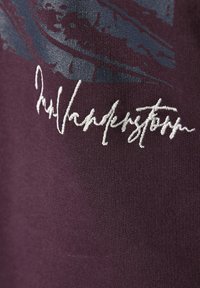 Maroon t-shirt fabric features a blue graphic design and white embroidered signature. The texture appears smooth with a slight sheen.