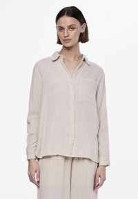 Camisa de manga larga beige claro hecha de una tela suave y texturizada. Cuenta con un cierre de botones en la parte delantera, cuello y un único bolsillo en el pecho.