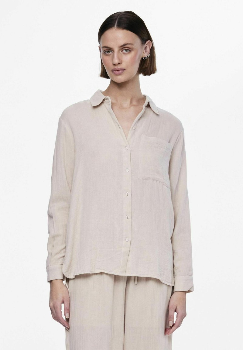 Camisa de manga larga beige claro hecha de una tela suave y texturizada. Cuenta con un cierre de botones en la parte delantera, cuello y un único bolsillo en el pecho.