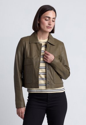 Vrouw met kort donker haar, gekleed in een olijfgroen jasje over een crème-zwart gestreept shirt en zwarte broek, voor een effen achtergrond.