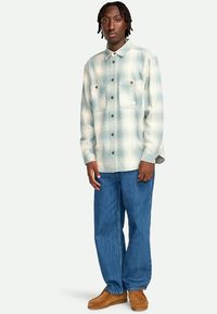 Mann mit geflochtenen Haaren, der ein hellblaues und weiß kariertes Hemd mit Knöpfen, lockere blaue Jeans und braune Wildlederschuhe trägt, steht vor einem weißen Hintergrund.