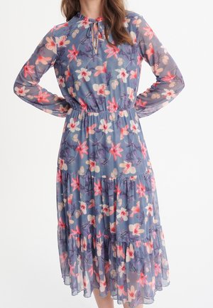 Femme portant une robe bleue à manches longues de longueur moyenne, avec un motif floral rose et blanc, un décolleté en forme de trou de serrure et une taille élastique, debout les mains sur les hanches.