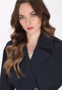 Tengerész stílusú trench coat széles gallérral, nagy gombokkal és sima anyagból készült. A modellen hullámos barna haj van, alatta pedig egy világos csipkeblúz látható.