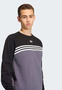 Čierna a sivá mikina s bielymi horizontálnymi pruhmi, raglánovými rukávmi a malým logom na hrudi. Mäkká textúra látky.