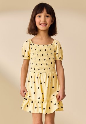 Lächelndes junges Mädchen mit schulterlangem dunklem Haar, das ein gelbes Kleid mit schwarzen Punkten und Puffärmeln trägt, steht vor einem beigen Hintergrund.