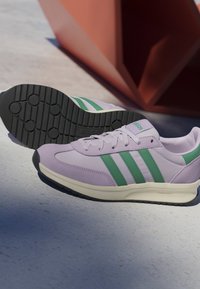Par de zapatillas Adidas de color lila claro con franjas verdes y suelas negras sobre una superficie gris claro cerca de una estructura angular naranja.