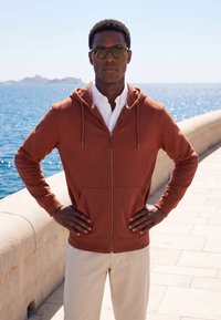 Homme portant des lunettes de soleil, un sweat à capuche zippé couleur rouille et un pantalon clair, debout les mains sur les hanches près d'une promenade en bord de mer sous un ciel dégagé.