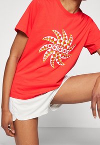 Röd bomulls-t-shirt med en geometrisk snurrdesign i vitt, gult och grönt, med "adidas"-logotypen centrerad i grafiken.