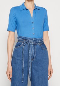 Blue short-sleeve polo shirt with a v-neckline and buttons, paired with high-waisted denim trousers featuring a tied belt and wide legs.