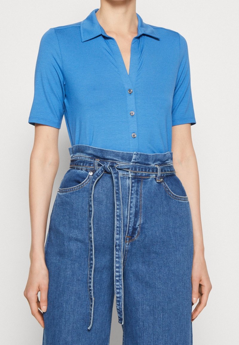 Blue short-sleeve polo shirt with a v-neckline and buttons, paired with high-waisted denim trousers featuring a tied belt and wide legs.