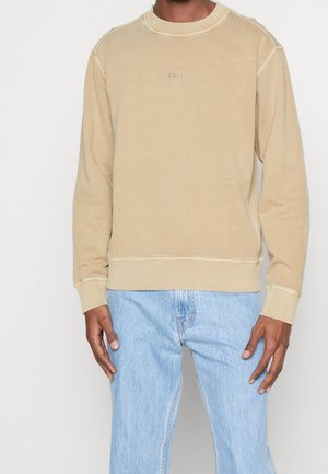 Mann trägt beige Sweatshirt mit langen Ärmeln und hellblaue Jeans, steht mit entspannten Armen an den Seiten.