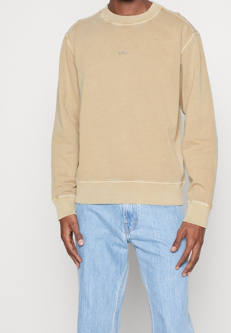 Homme portant un sweat-shirt beige à manches longues avec col rond et un jean en denim bleu clair, debout, les bras détendus le long du corps.