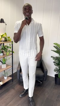 Weißes Hemd mit senkrechten Streifen, kombiniert mit schmalen weißen Hosen und schwarzen Slippern. Steht vor einem Pflanzenregal.
