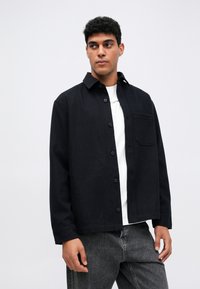 Schwarze button-up Hemdjacke aus strukturiertem Stoff, mit einer einzelnen Brusttasche und einem Kragen, getragen über einem weißen T-Shirt.