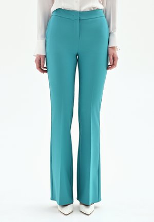 Femme portant un pantalon évasé taille haute bleu sarcelle associé à un chemisier blanc à manches longues et des escarpins blancs à bout pointu, debout devant un fond blanc.